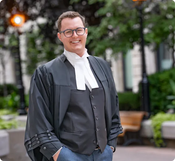 Smiling young man wearing glasses, a black barrister robe, and white neckbands standing outdoors with hands in pockets.
